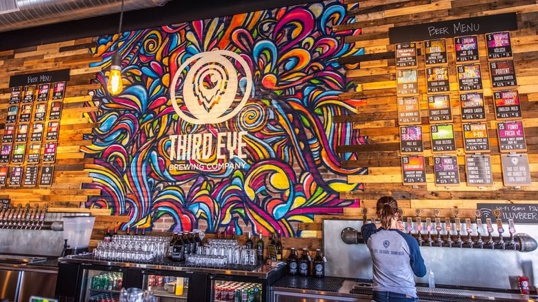 Colorful mural on wooden wall behind bar counter at Third Eye Brewing, with beer menu and bartender pulling a beer from tap