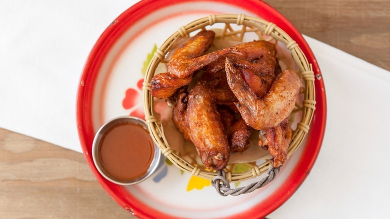 Bamboo basket of chicken wings next to sauce on a decorative white plate with flowers and a red rim