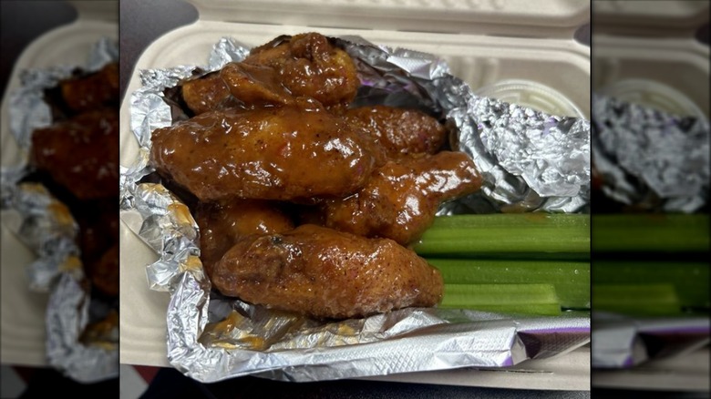 Saucy chicken wings in a tin foil lined to go box next to celery sticks and ranch