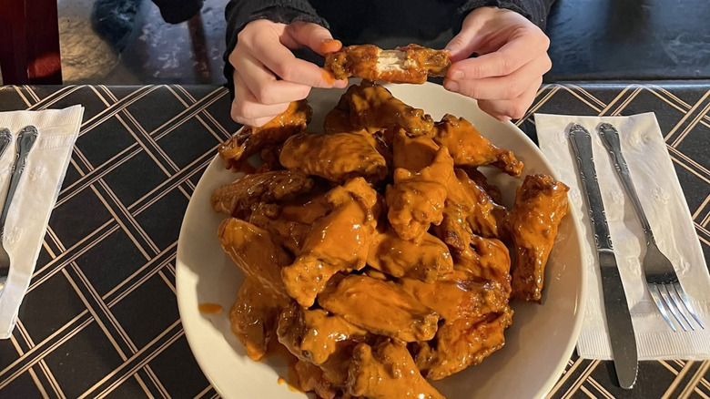 Two hands holding a chicken wing above a massive pile of Dirt Wings on a white plate