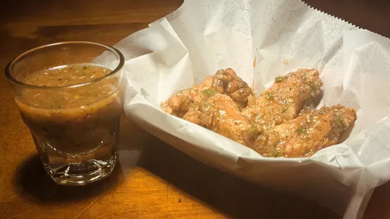 Chicken wings in a paper bowl next to a small glass of sauce
