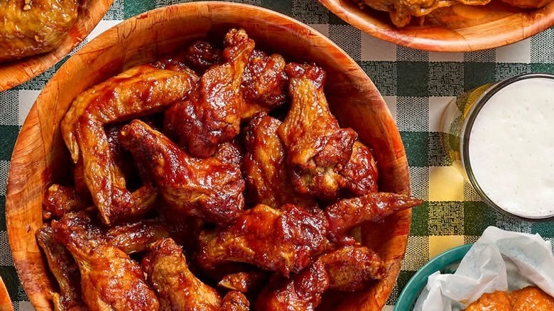 Wooden bowl of saucy chicken wings on a table next to three other wing bowls and a glass of beer