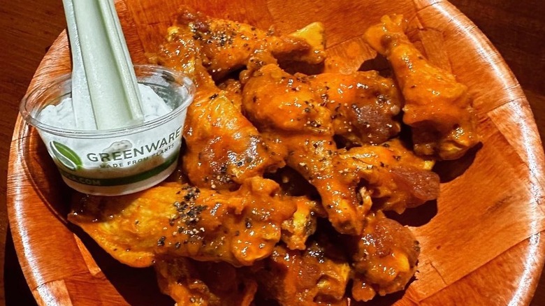 Wooden bowl of saucy and peppery wings next to plastic container of dipping sauce