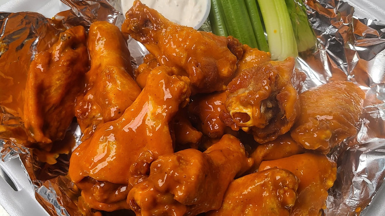 Styrofoam container of saucy buffalo wings on top of aluminum foil with celery sticks and blue cheese