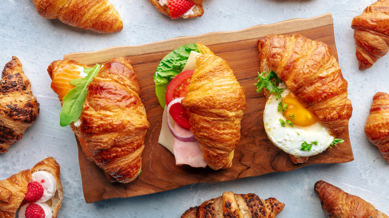 Croissants sandwiches with various fillings on wooden serving board surrounded by more croissants
