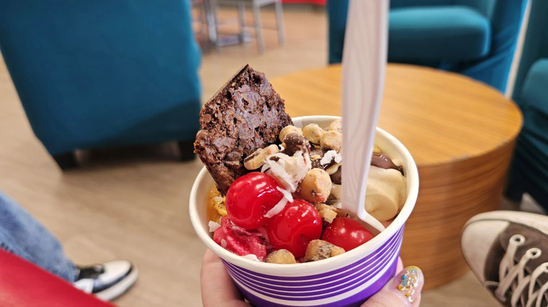 hand holding a purple cup of froyp with cherries, cookie dough pieces, brownie, and spoon