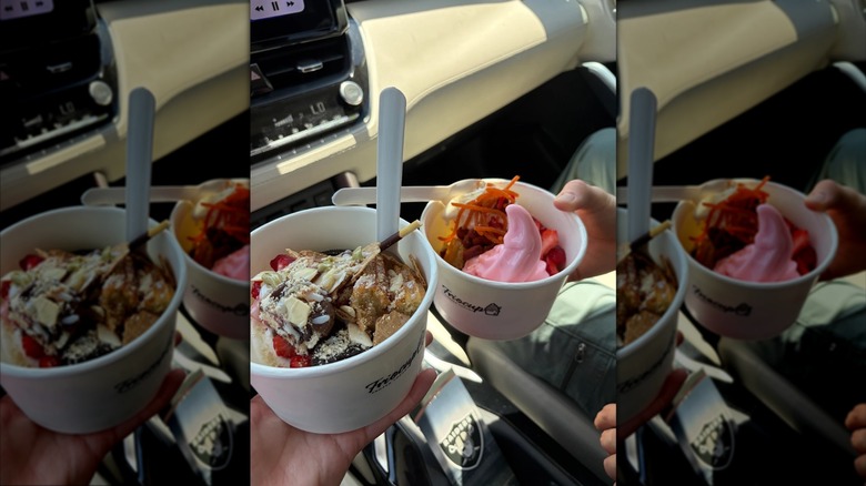 two hands holding two paper cups of froyo in a car with a variety of different toppings