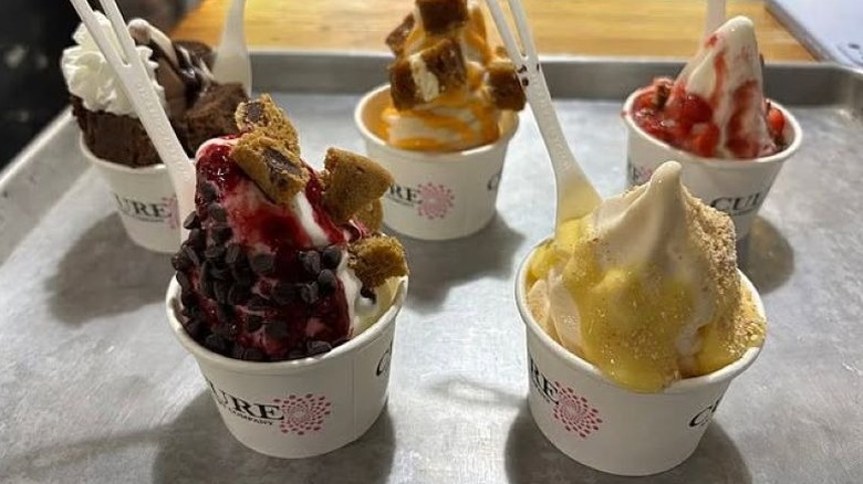 six cups of froyo with a variety of different toppings and fruit drizzles on a metal tray