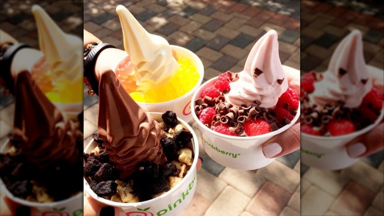 hands holding three cups of froyo swirls with a variety of toppings like fruit, cookies, and boba