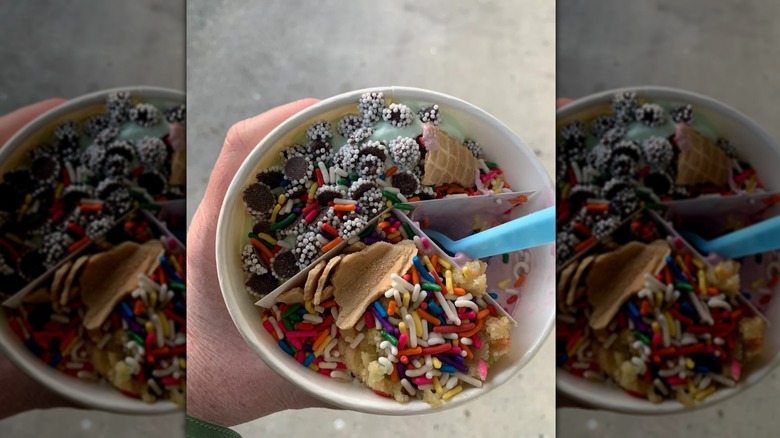 hand holding a paper cup of froyo with a divider and three different flavors with a variety of different toppings and blue plastic spoon