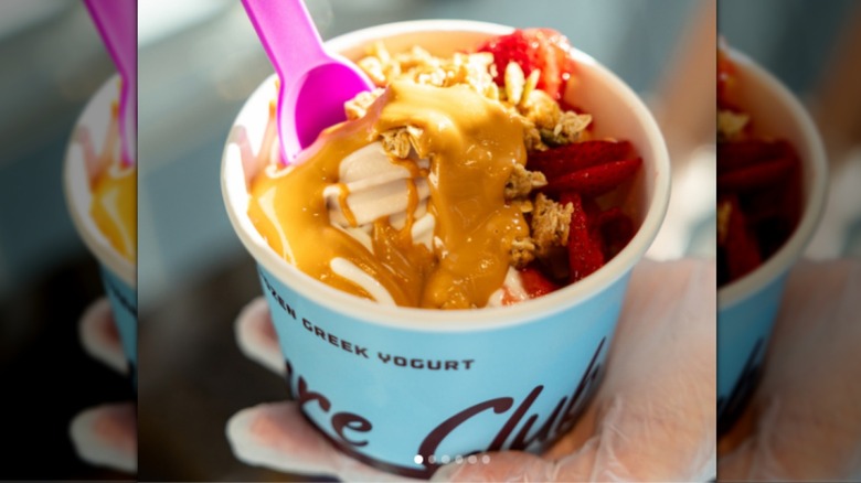 cup of froyo with caramel syrup, granola, fresh strawberry chunks, and a pink plastic spoon