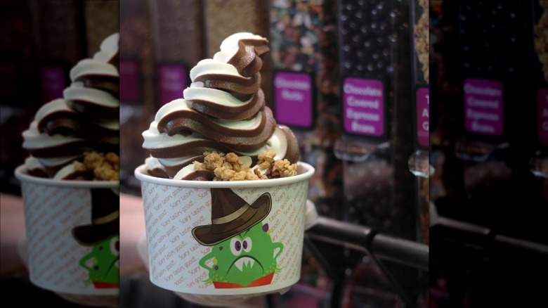 cup of brown and white froyo swirl with granola with an array of toppings in the background