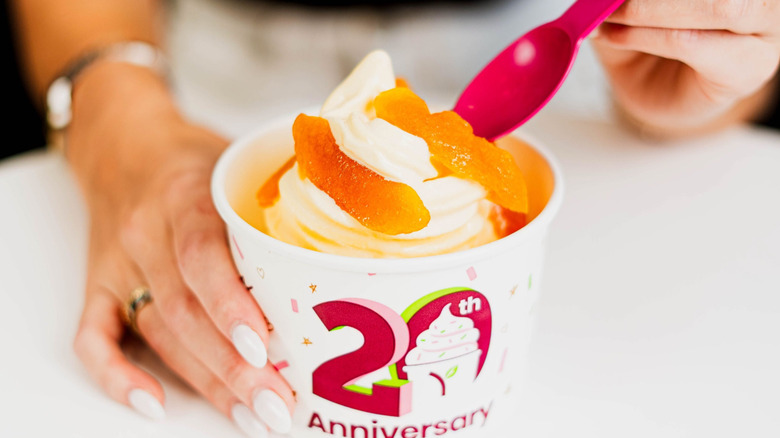 hand with white nails dipping a pink plastic spoon into a curl of froyo with orange gummy slice toppings