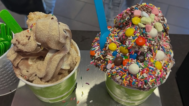 two cups of froyo piled high with an enormous amount of toppings like m&ms, sprinkles, and marshmallows with green and blue plastic spoons