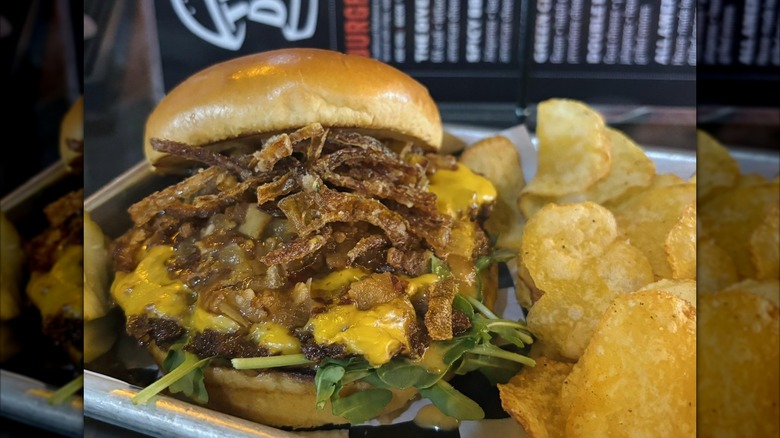 Closeup of The Crispy Jam Burger at The Dive piled high with cheese and toppings and a side of potato chips all on metal tray