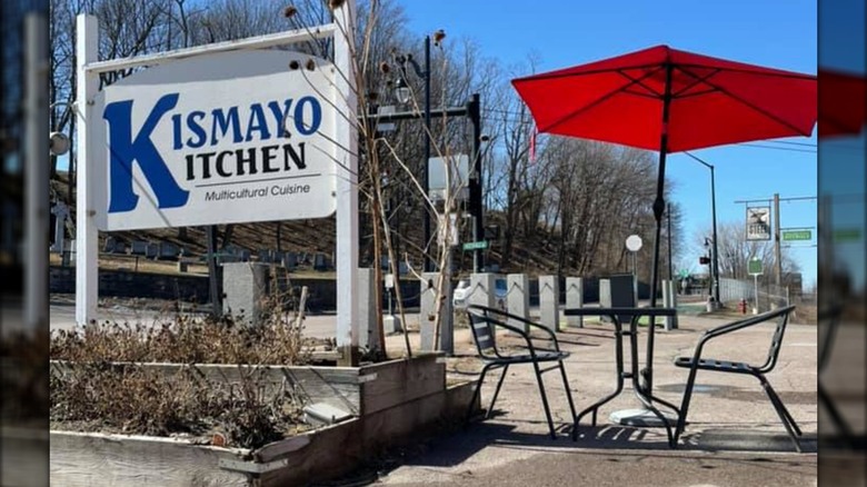 Exterior of Kismayo Kitchenn on sidewalk with white restaurant sign and small picnic table with red umbrella