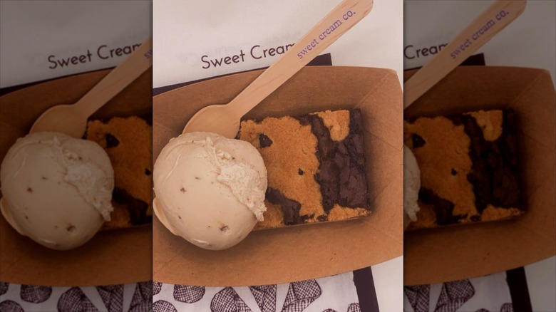 Ice cream scoop and a cookie brownie in a cardboard tub with a wooden spoon