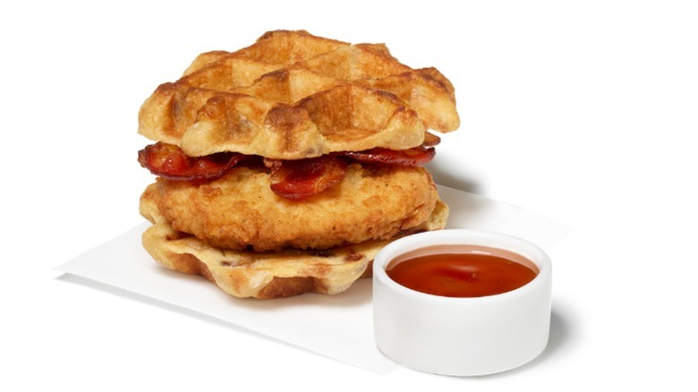 Chick-fil-A Chicken & Waffles Breakfast Sandwich with a cup of dipping sauce on a white background.