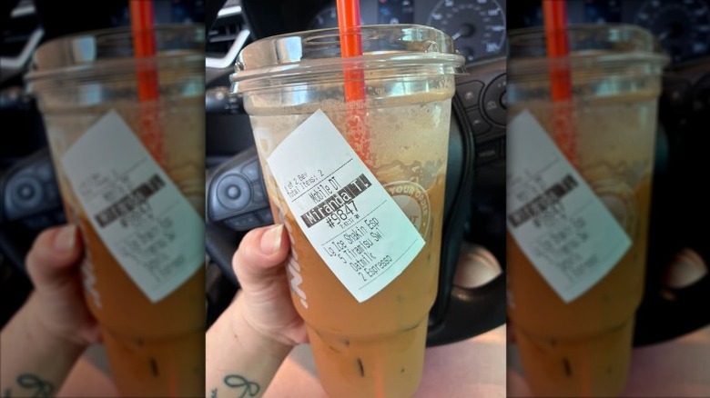 A hand holding a Dunkin' Tiramisu Shakin' Espresso in a plastic cup in front of a car dashboard