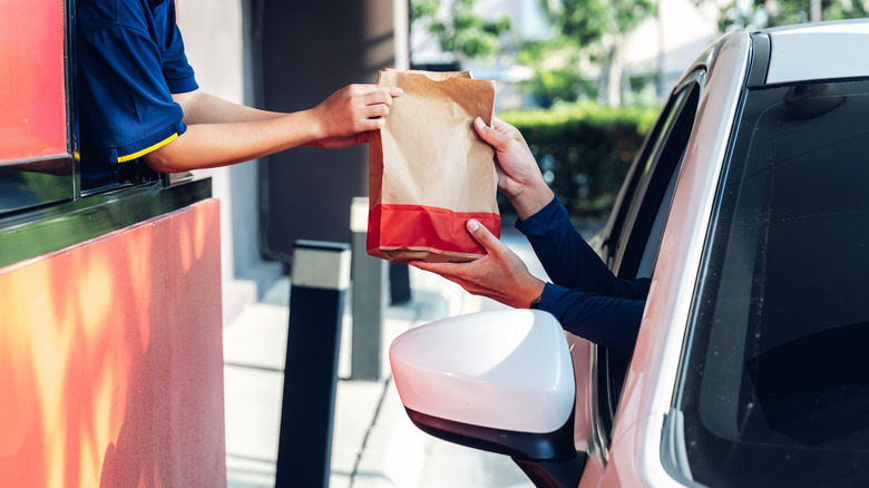 A drive-thru worker handing a brown and red paper bag to a person in a white car