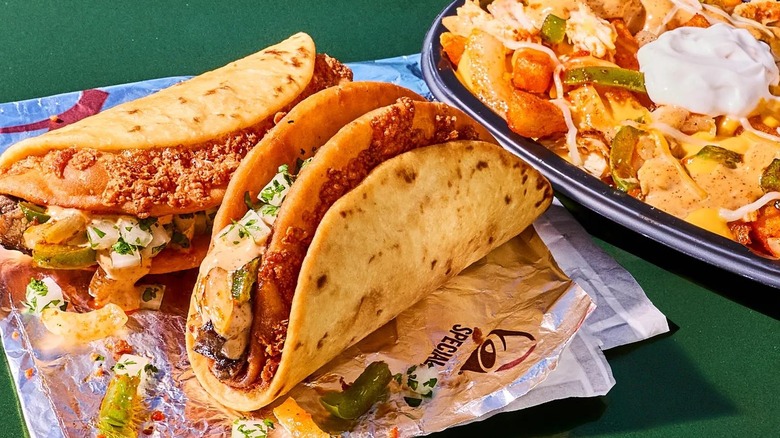 Two Fajita Street Chalupas on Taco Bell packaging atop a green surface with a black plastic bowl of loaded fries beside them