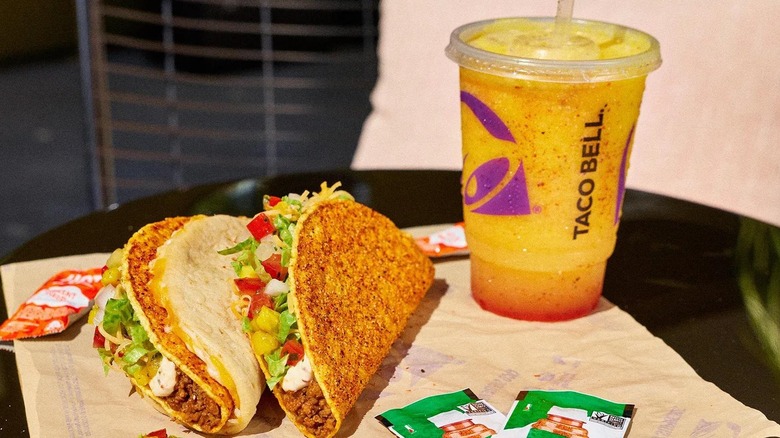 Two tacos and a yellow and red frozen drink in a Taco Bell cup sitting on a tray, with two small Tajin packets in the foreground
