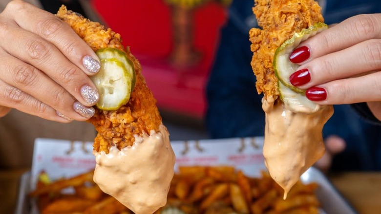 Two hands shown, each holding a Nashville Hot Chicken tender strip with pickles and dipping sauce at Angry Chickz