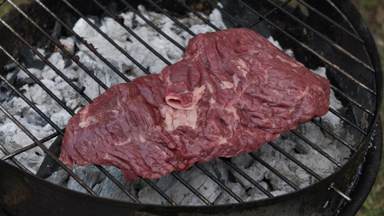 Raw flank steak being cooked over hot charcoal on a grill