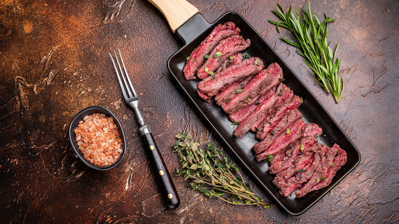 Sliced cooked flank steak in pan alongside a meat fork, sprigs of rosemary and oregano, and a small bowl of pink sald
