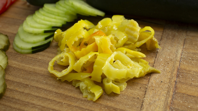 Pile of sliced banana peppers next to sliced cucumbers and pickles on wooden table