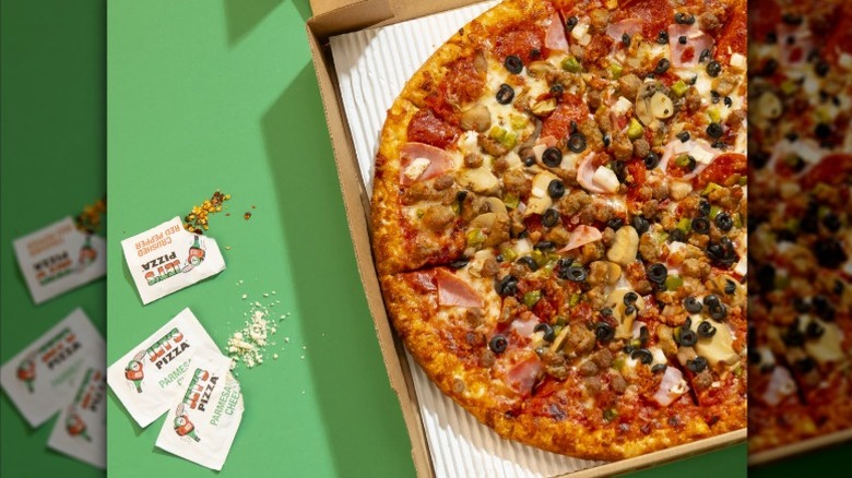 Jet's Pizza Jet 10 pizza with meat and vegetables in a cardboard delivery box with packets of parmesan cheese on a green surface.