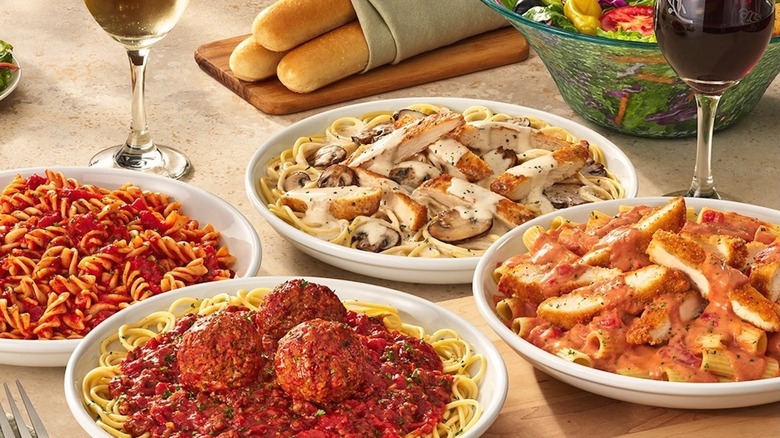 An assortment of Olive Garden pastas on a table with wine, salad, and bread sticks.