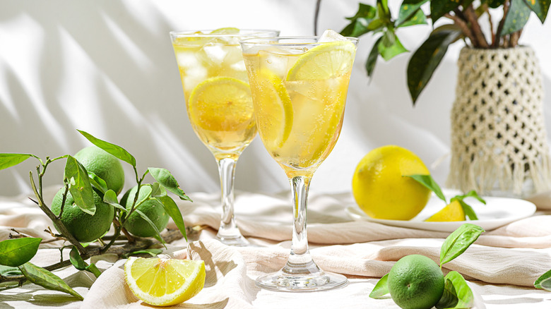 Two stemmed glasses with lemon water surrounded by lemons and limes.