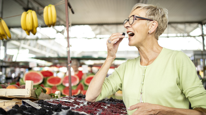 Elderly woman sampling blackberry at market with other fruit and berries in the background