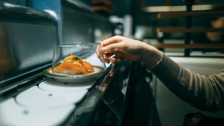 Someone reaching out to pick up a plate of sushi from a conveyor belt