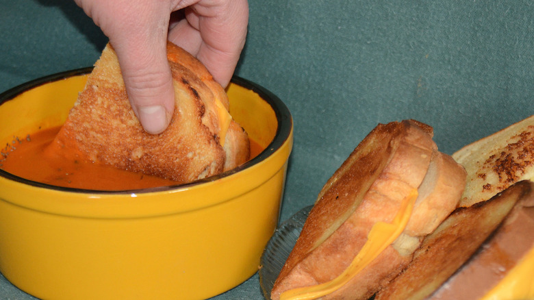 A hand dunks half of a grilled cheese sandwich inside a yellow bowl of tomato soup