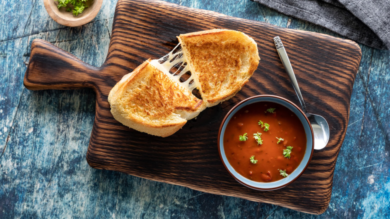 Top-down view of a sliced grilled cheese sandwich with a generous cheese-pull sits next to a cup of tomato soup on a wooden cutting board