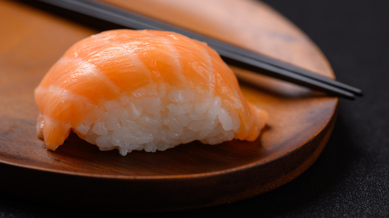 A piece of salmon nigiri on a wooden plate with two black chopsticks behind it