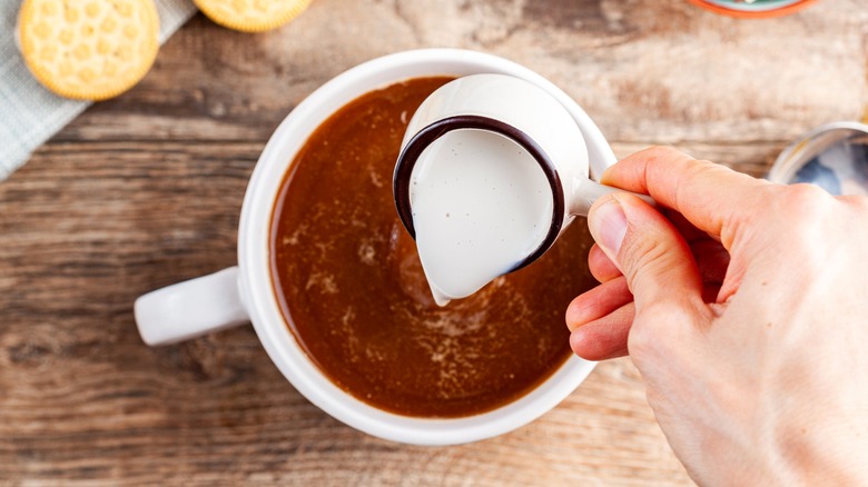 Person pouring creamer into white cup of coffee