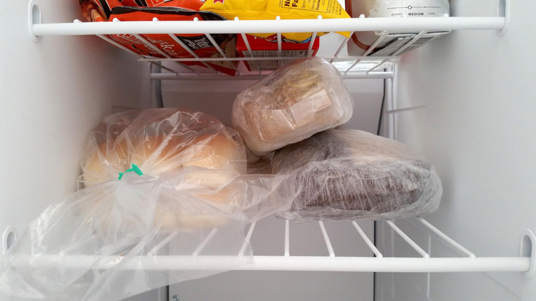 Loaves of bread wrapped in plastic and foil stored on a freezer shelf alongside frozen packaged goods.