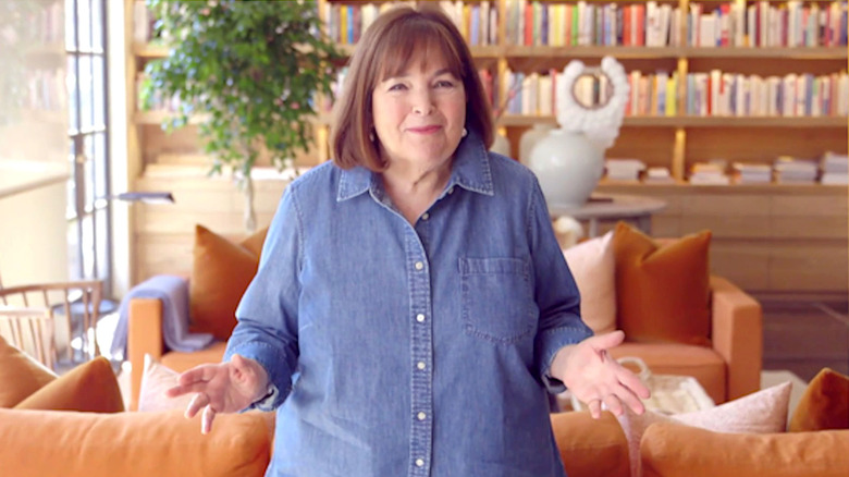 Ina Garten standing in a cozy living room wearing a denim button-down shirt and smiling with hands raised, a bookshelf and orange couch in the background