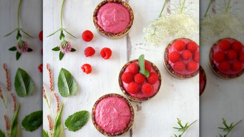 Pink raspberry tarts with a brown crust in mason jar lids on a white surface with scattered berries and herb leaves