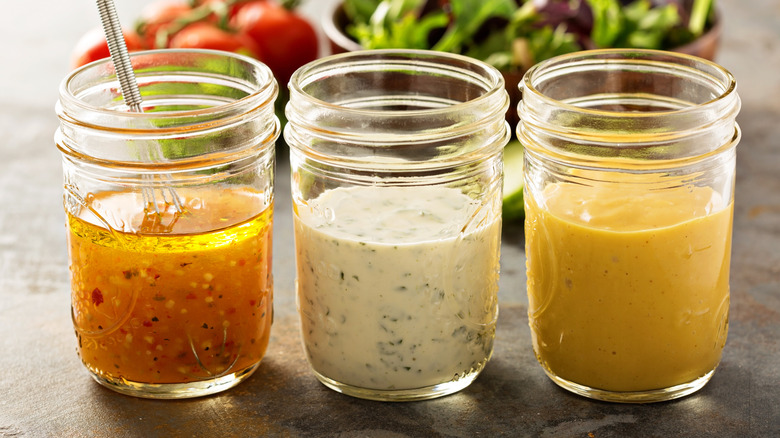 Three glass jars of salad dressing with bowl of lettuce and tomatoes in the background