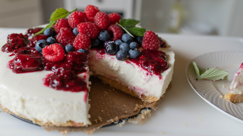 A no-bake cheesecake topped with a berry sauce, fresh berries, and mint, with one sliced cut out of it