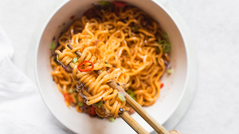 Spicy instant noodles with slices of green and red chili with chopsticks in a white ceramic bowl.
