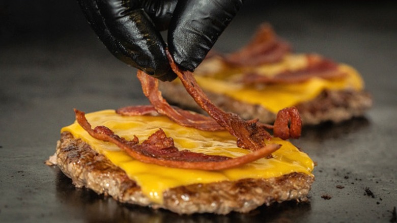 A gloved hand arranging bacon on a cheeseburger patty on a grill.