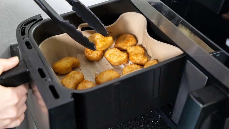 hands using tongs to hold a chicken nugget above an air fryer basket
