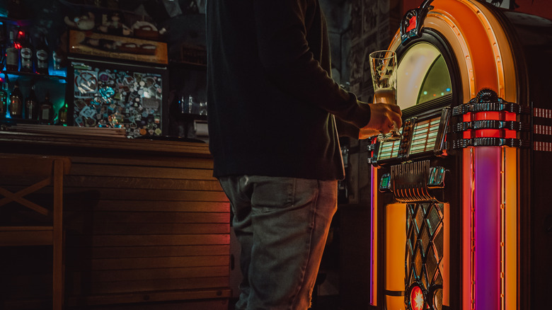 A person holds a beer and selects songs on a jukebox in a bar