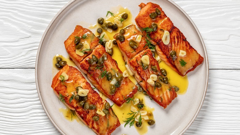 Four rectangular pieces of crispy browned salmon on a yellow piccata sauce with garlic and capers on a round white plate