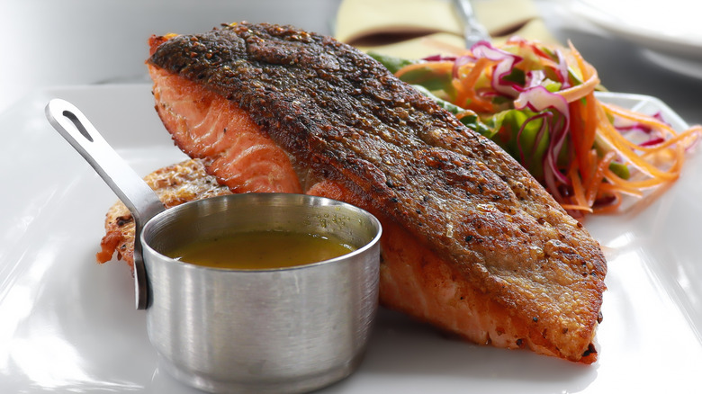 Crispy cooked salmon propped up on a white plate with a small bowl of a yellow sauce in front and shredded vegetables
